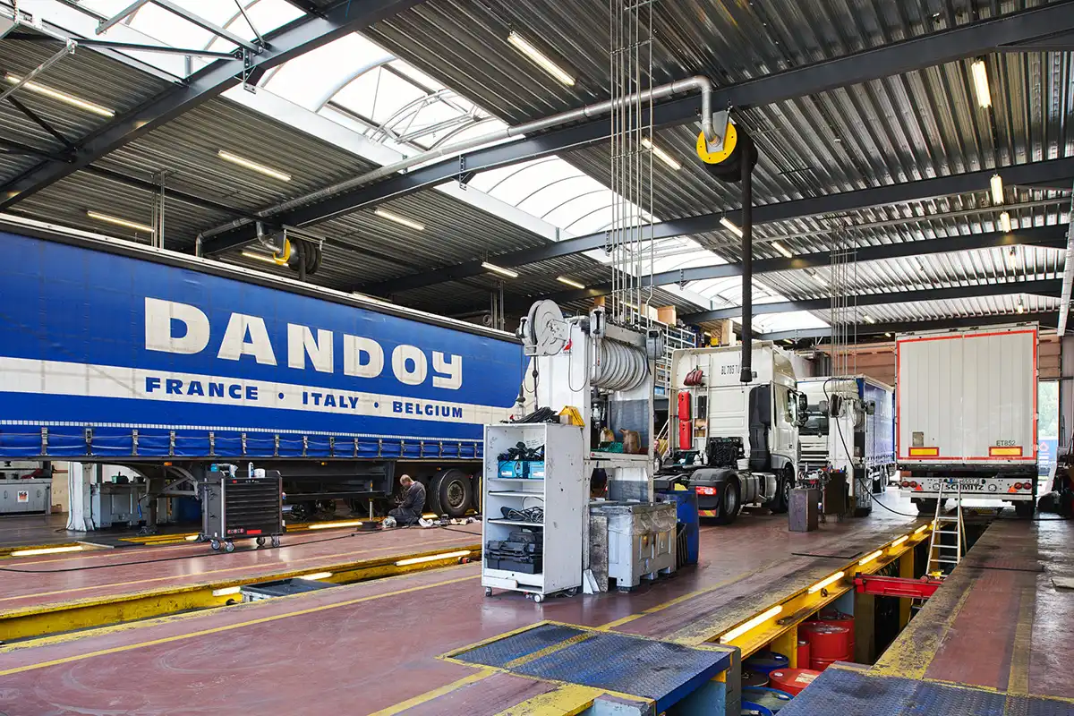 Camion bleu Dandoy dans un atelier, entouré d'équipements de maintenance, avec un autre camion blanc à l'arrière-plan.