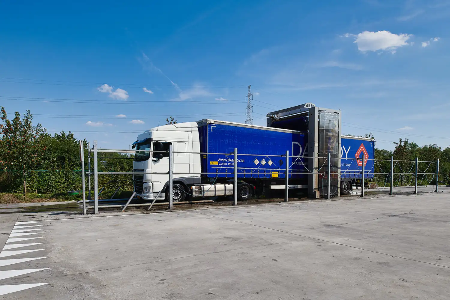 Un camion blanc et bleu marqué Dandoy est stationné dans une aire de lavage entourée de végétation.
