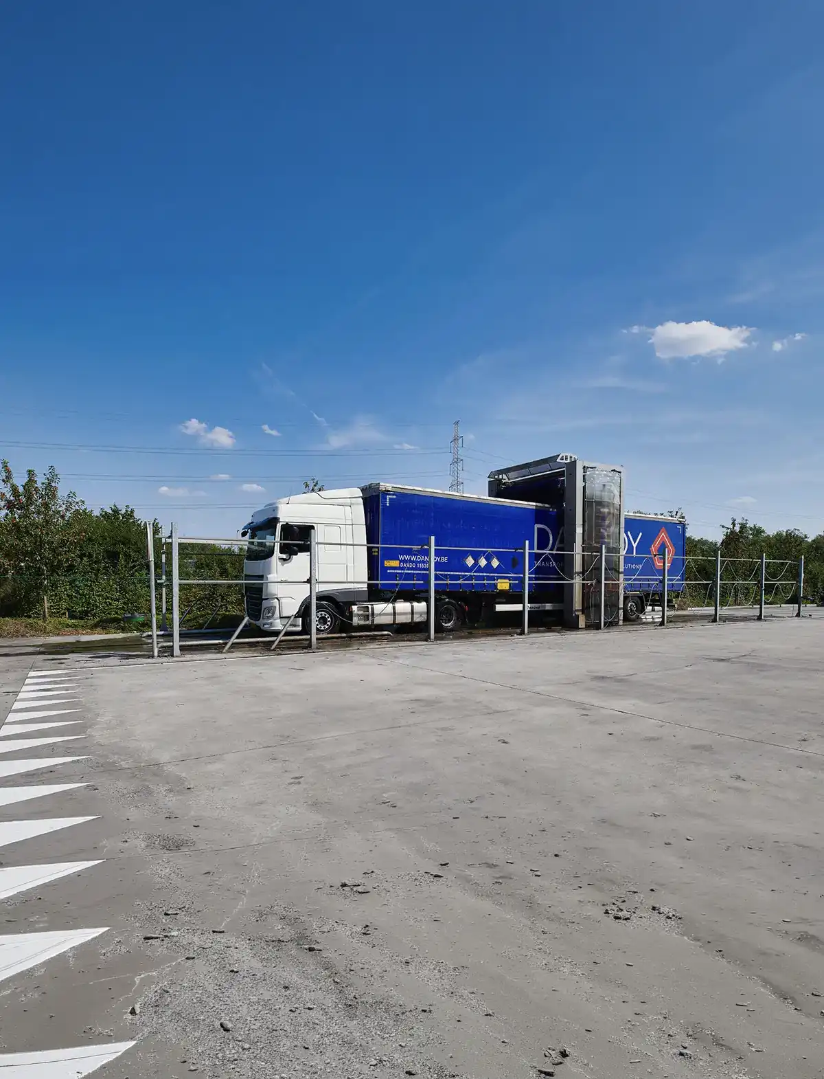 Camion blanc avec remorque bleue Dandoy garé sur un parking bétonné, ciel bleu en arrière-plan.