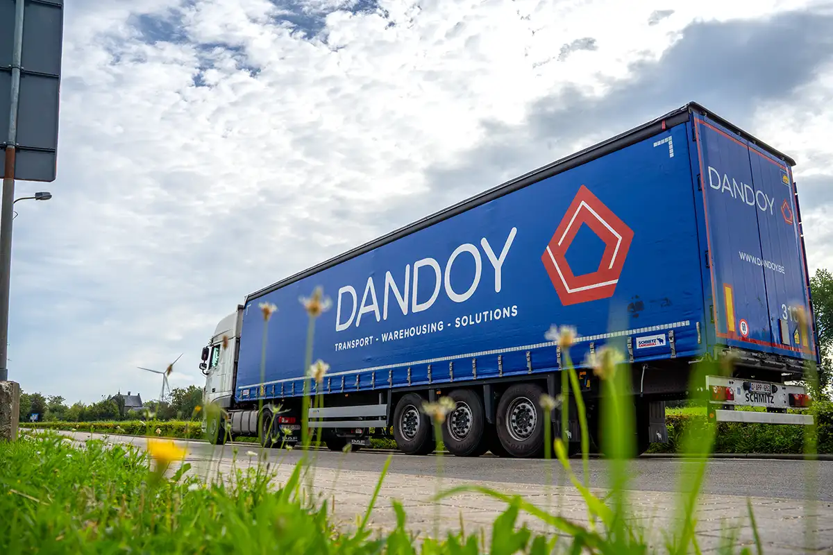 Un camion bleu de Dandoy Transports sur une route, avec un ciel nuageux et de l'herbe au premier plan.