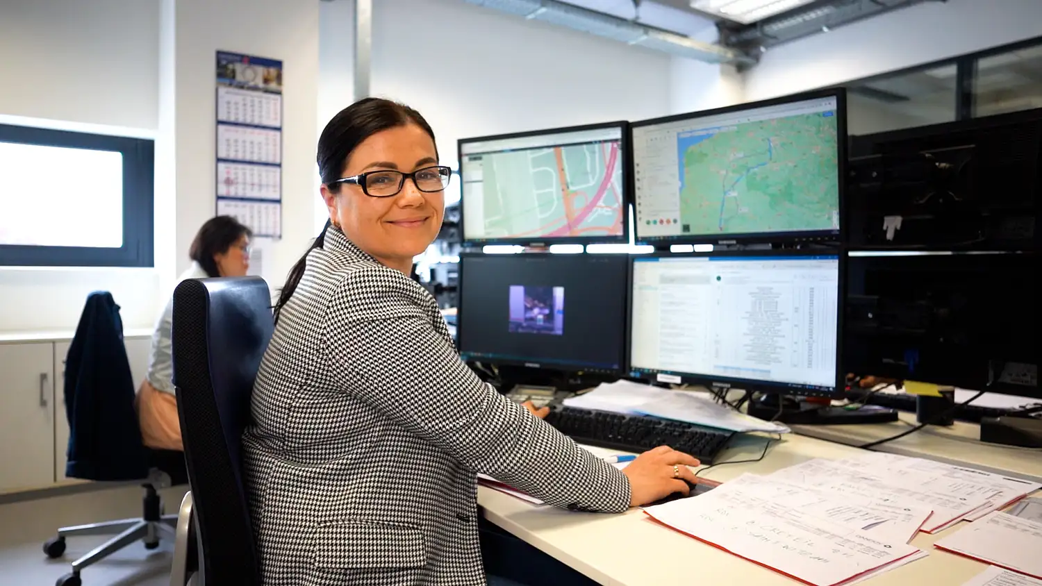 Une femme souriante travaille à un bureau avec plusieurs écrans d'ordinateur affichant des cartes et des documents, illustrant Dandoy 23.