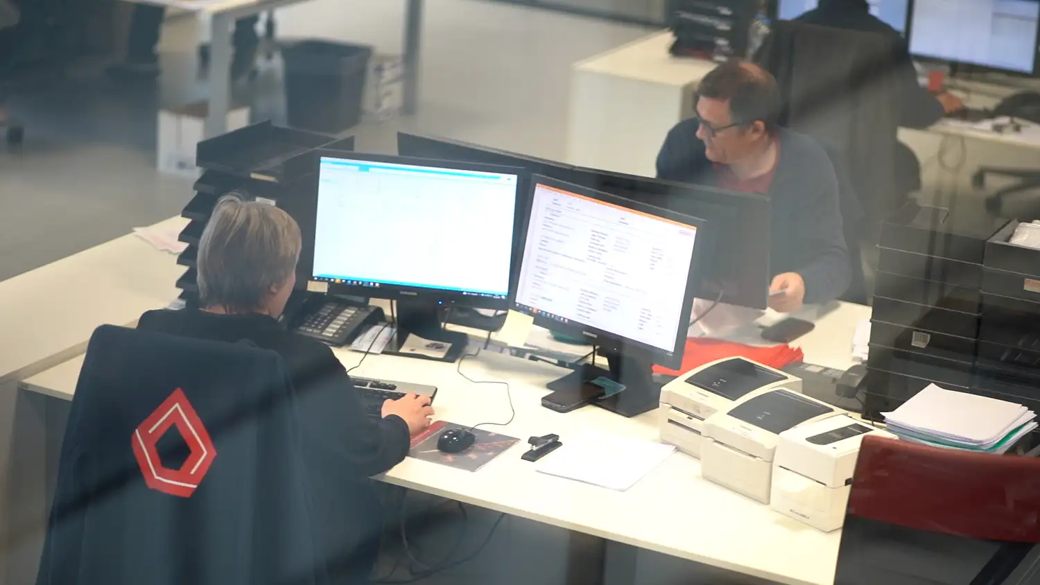 Deux personnes travaillent à des bureaux avec des écrans d'ordinateur et des imprimantes, dans un environnement de bureau moderne.