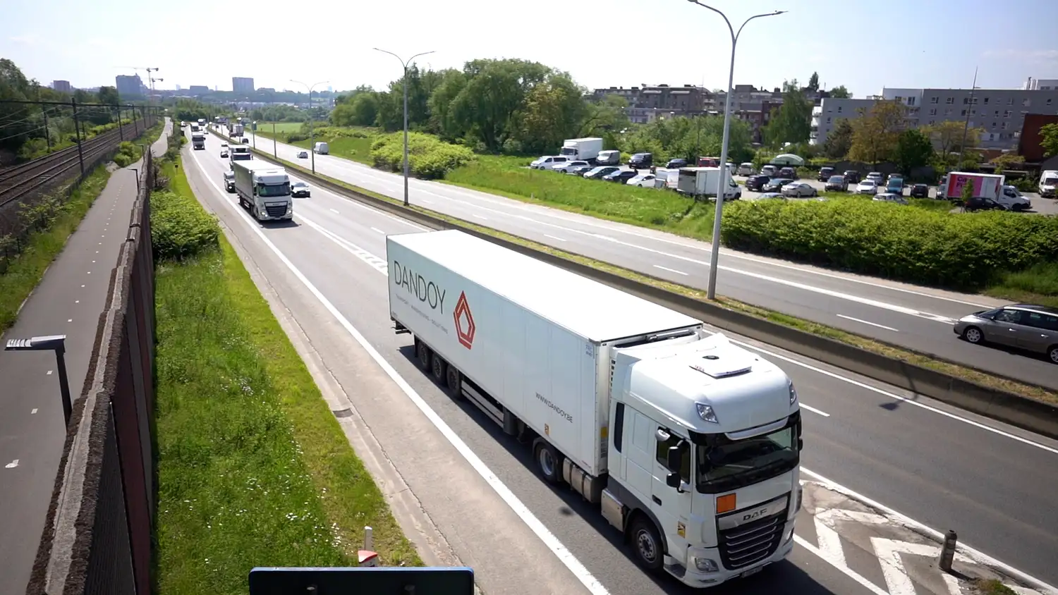 Camion blanc avec logo Dandoy sur une autoroute, entouré de verdure et de voitures, sous un ciel dégagé.
