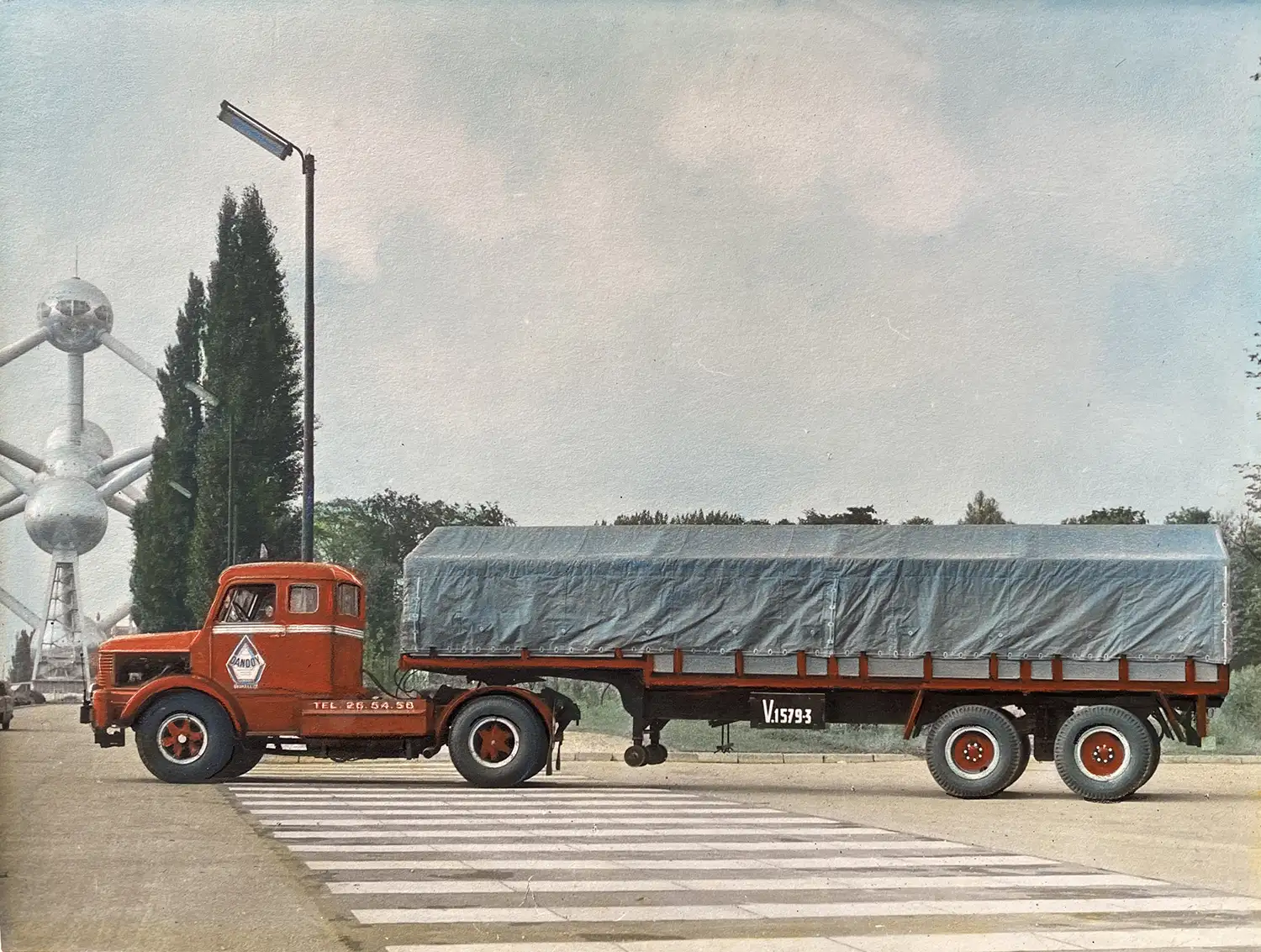 Camion rouge avec remorque couverte en toile bleue, stationné devant l'Atomium à Bruxelles, sur une route bordée d'arbres.