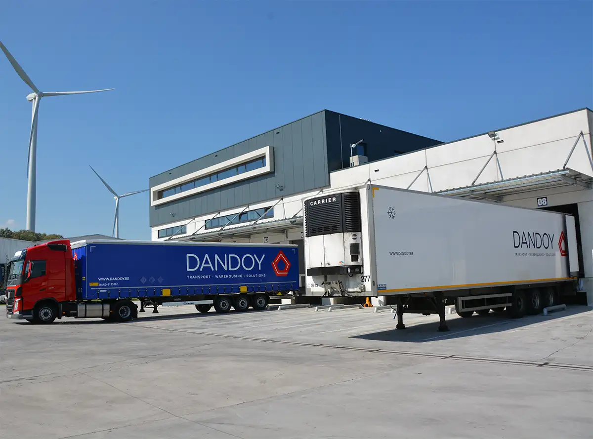 Deux camions de transport Dandoy sont stationnés devant un bâtiment industriel avec des éoliennes en arrière-plan, illustrant le service client en transport.
