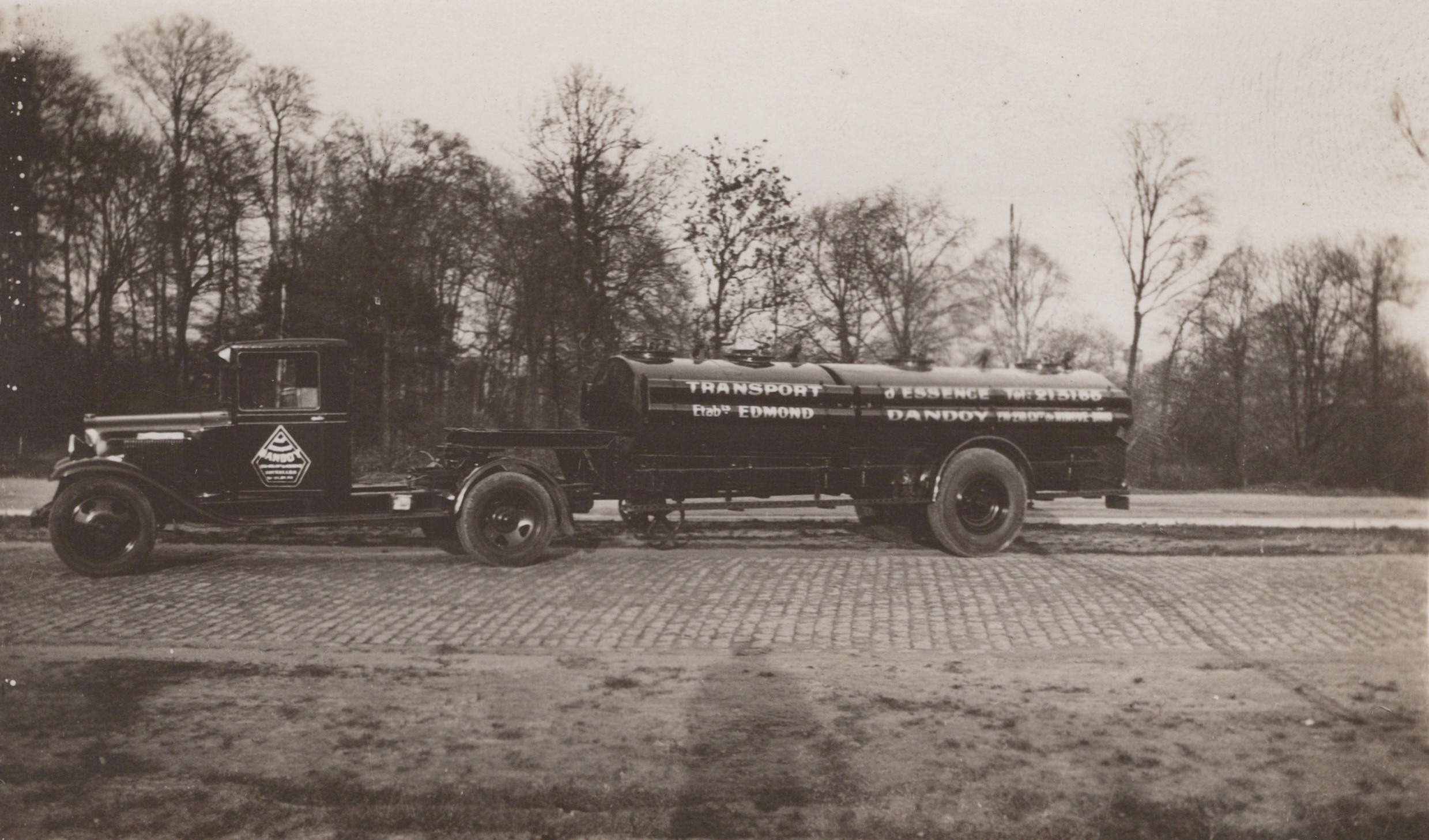 Camion-citerne noir "Transport d'essence Dandoy" stationné sur route pavée, arbres nus en arrière-plan, Photo 0041.