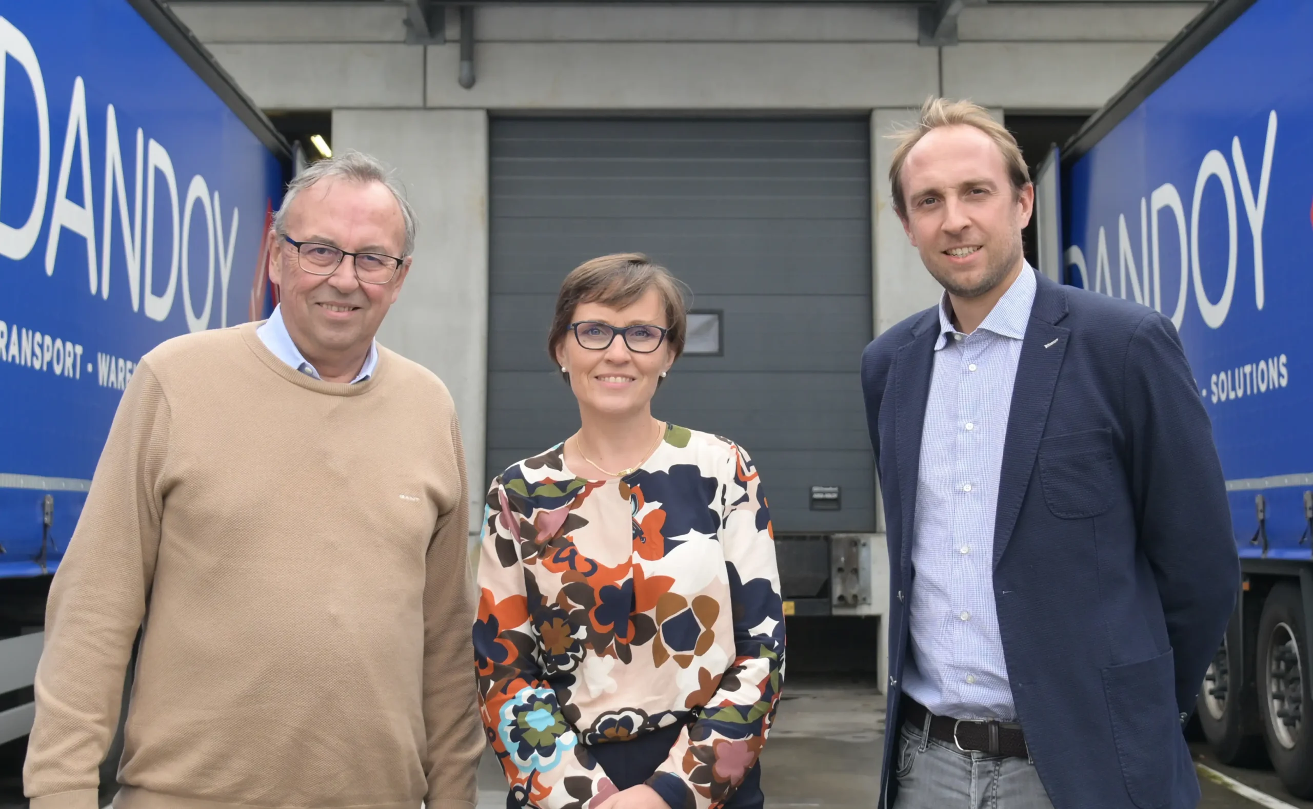 Trois personnes posent devant un entrepôt avec des camions bleus marqués "Dandoy", symbolisant la quatrième génération de l'entreprise.